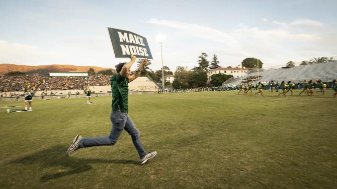 student running on the football field with a sign saying "make noise." 