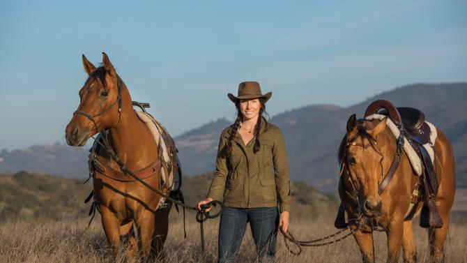 A woman stands between two brown horses and holds their reins in a field with mountains in the background.