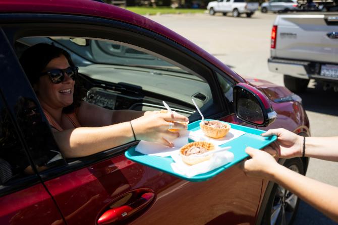 A smiling person in a red car grabs an ice cream in a waffle cup off of a blue tray.