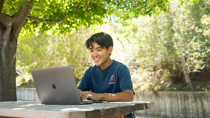 A student uses a laptop under tree canopy on campus
