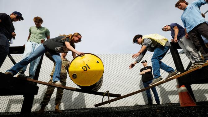A team of students move a yellow barrel across wooden planks