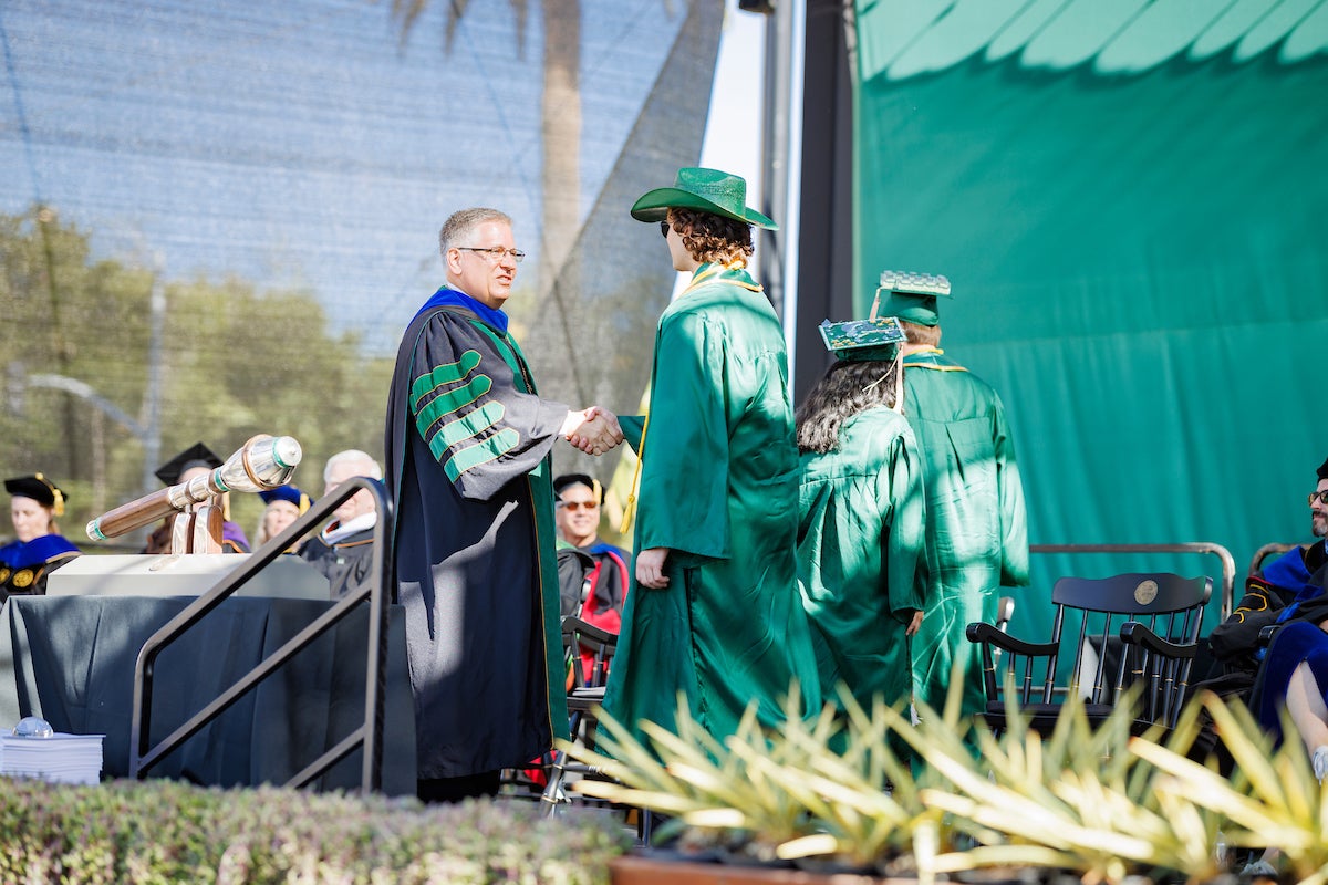 President Armstrong shakes hands with a graduate in a green cowboy hat at commencement.