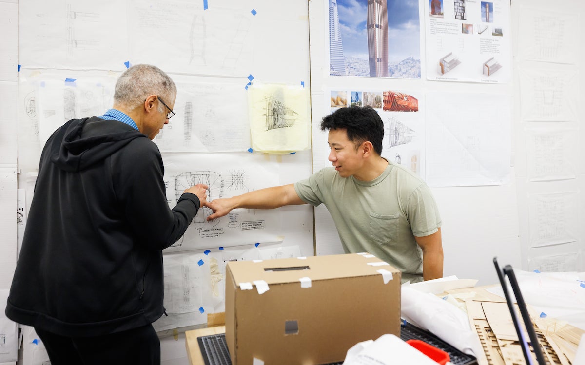 A professor and a student point to a set of drawings on a wall of an archtiecture studio.