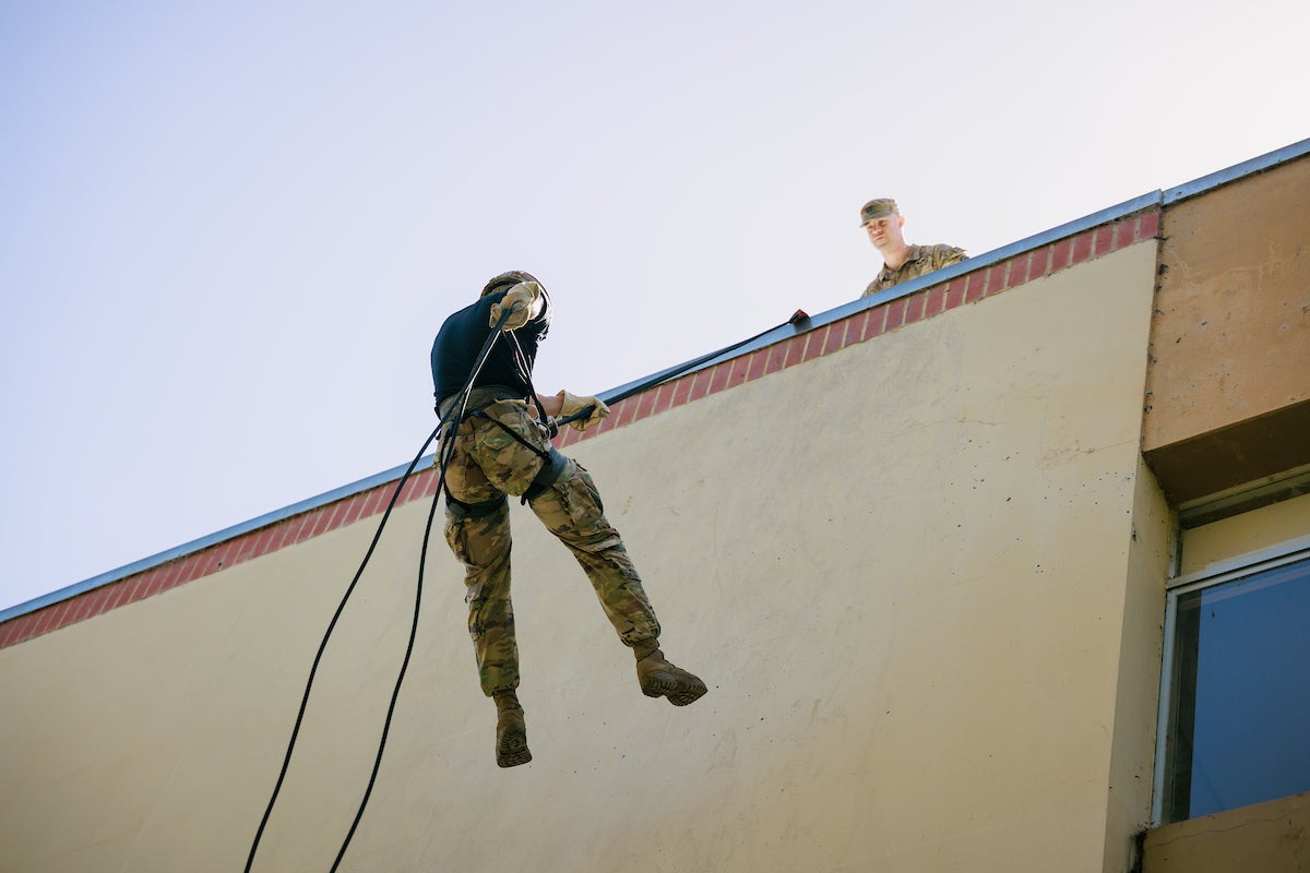 A student in camo pants rappells down the side of a building.