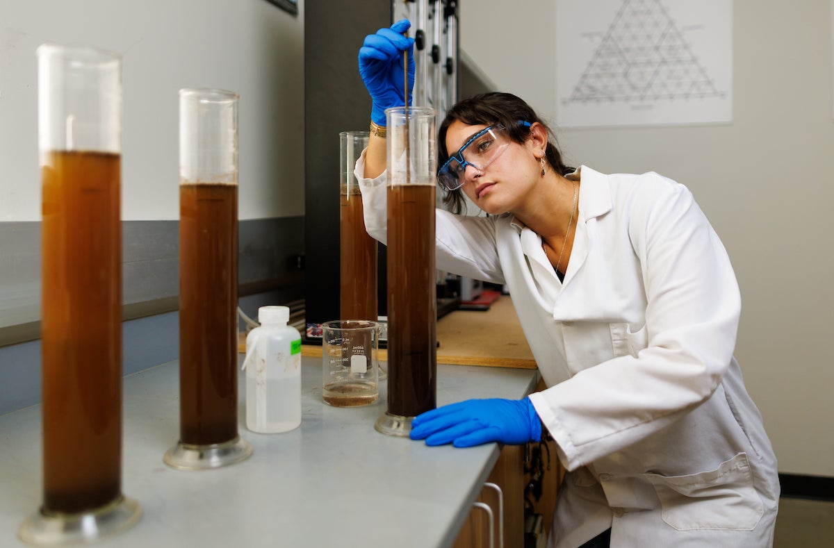 Nabila Wildlman wears a lab coat and protective gear as they stick a thin rod into a large beaker full of soil and water.
