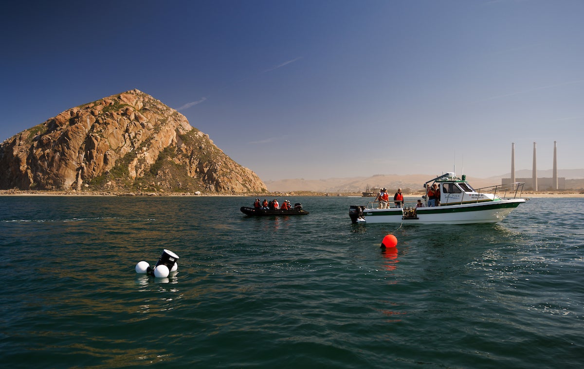 People in a black dinghy and a white research vessel in Morro Bay, testing an ocean powered generator in the water.