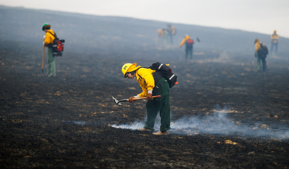 A student in firefighting gear leans over a steaming pocket of burned earth.