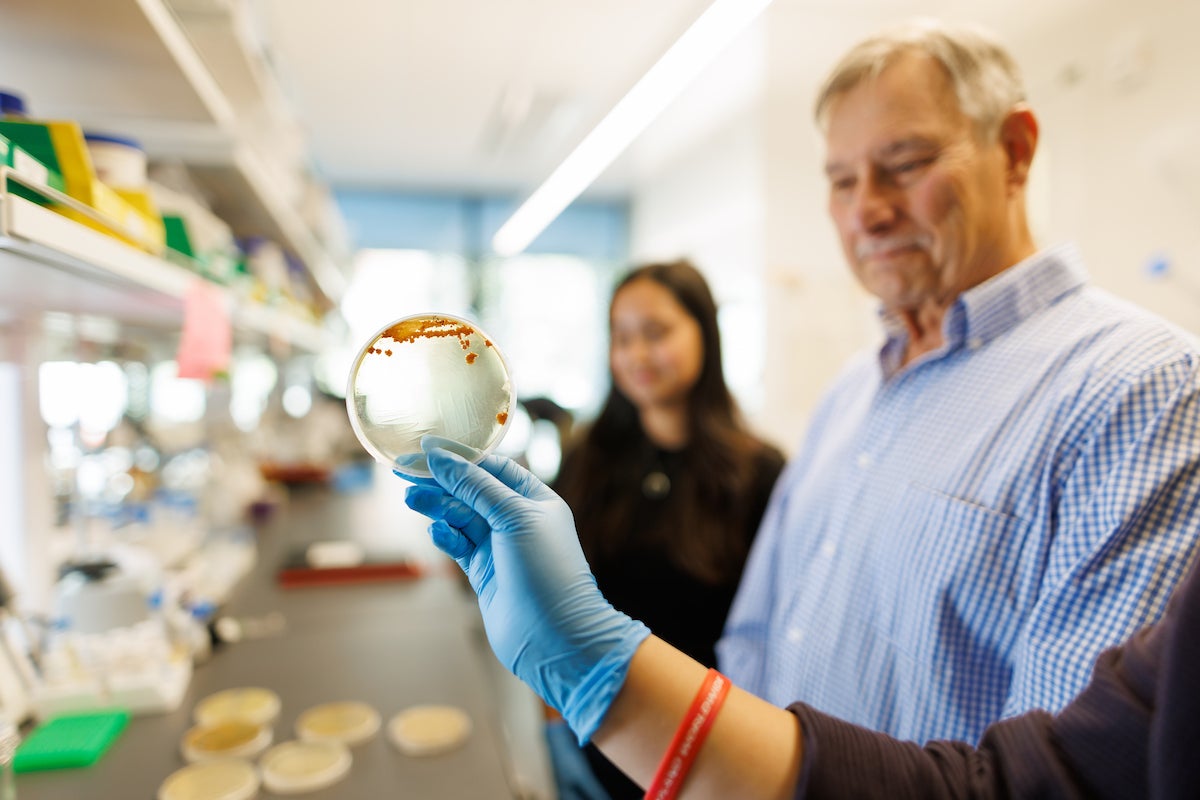 A hand holds a petri dish in front of a professor's face.