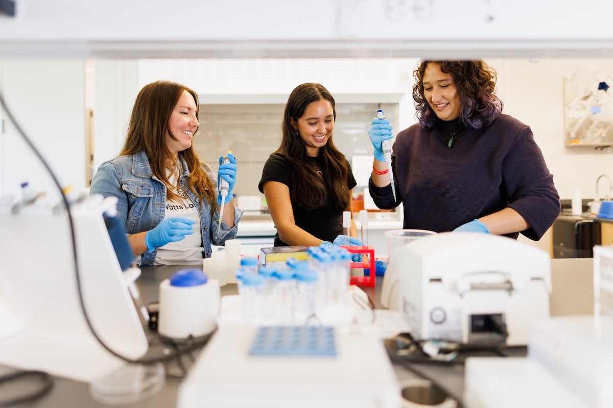 Three women hold droppers and work with scientific equipment.