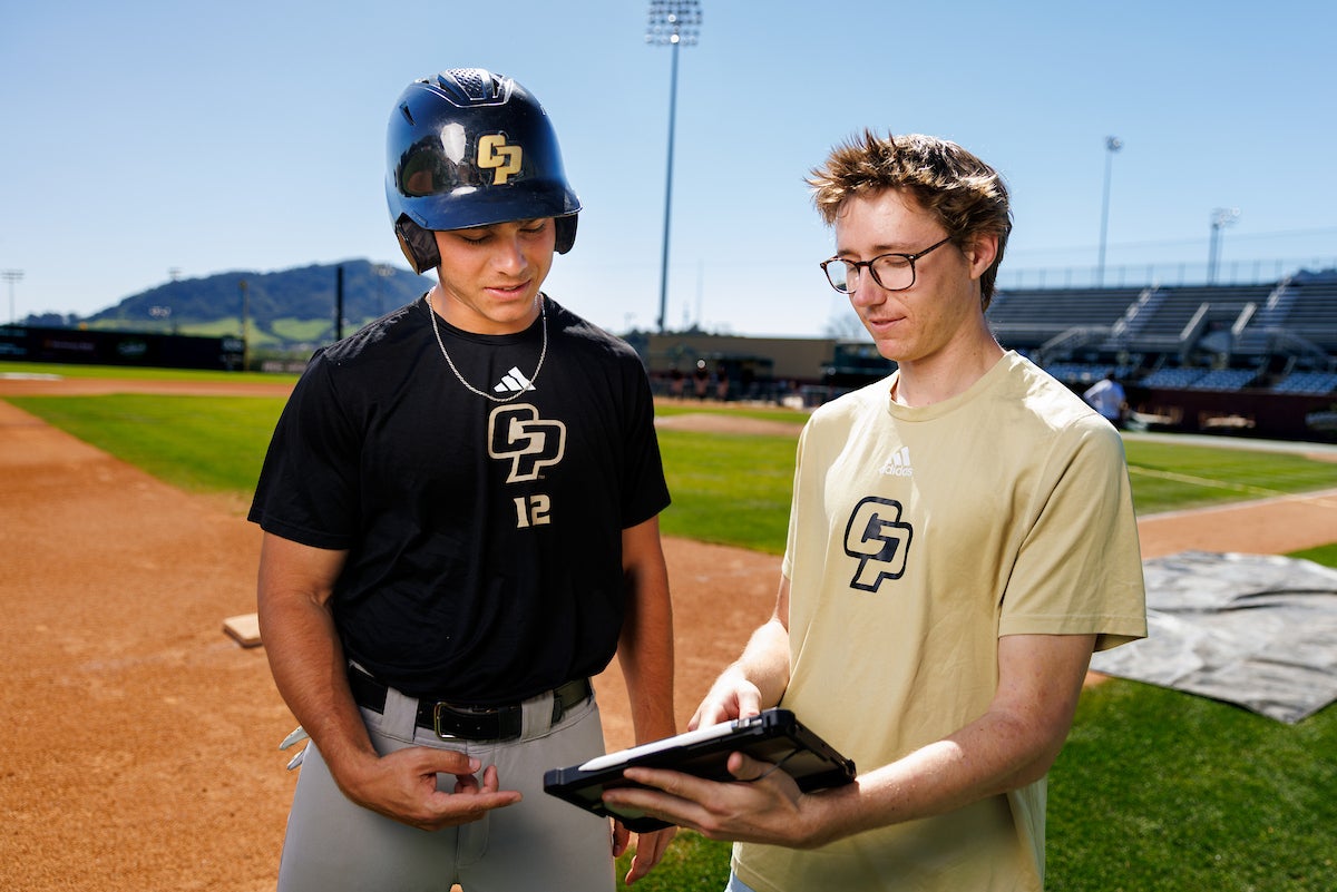 A Cal Poly baseball player on the left consults with Gabe Riedel on the baseball field.
