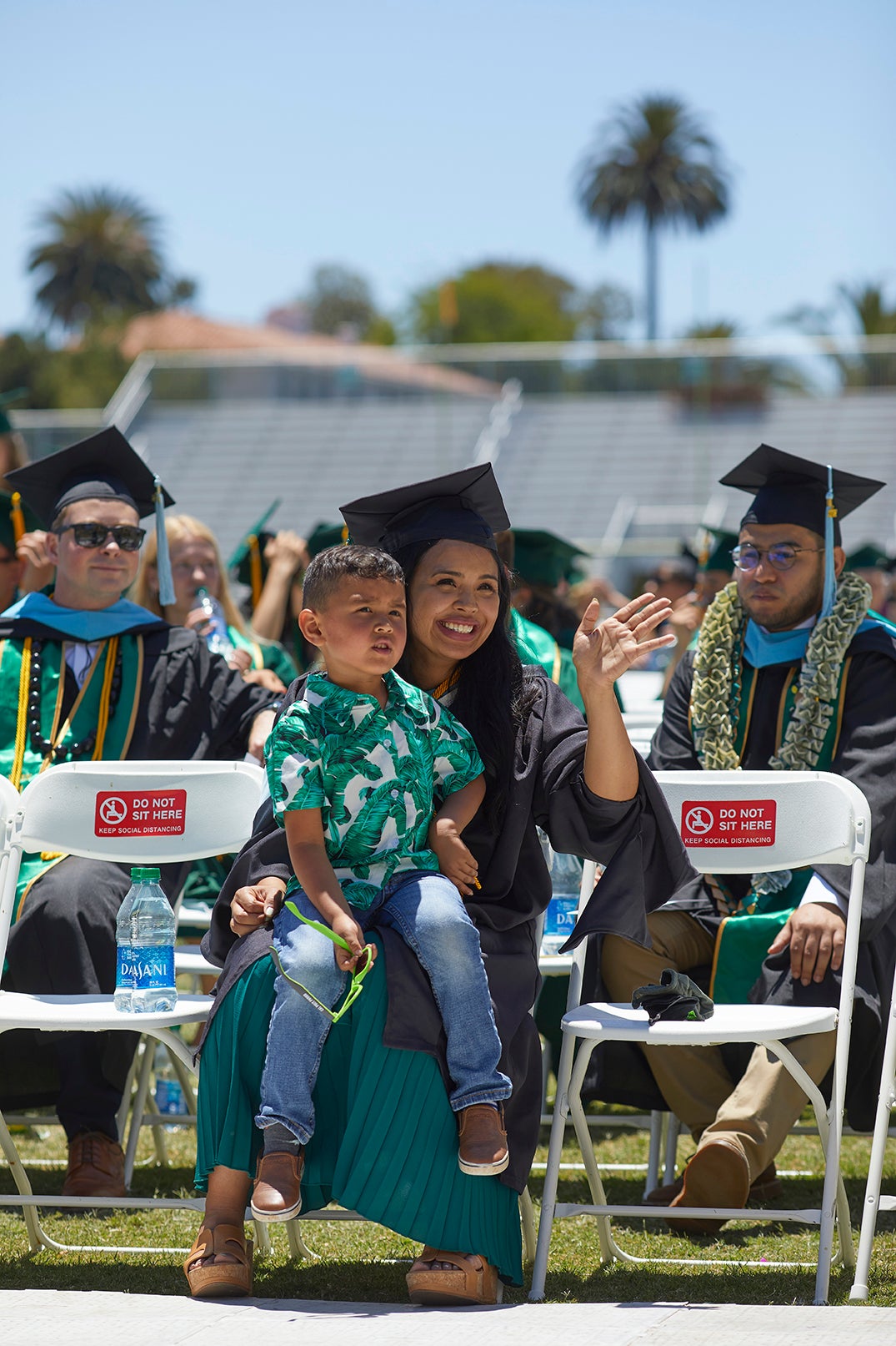 A graduating student smiles as she sits with fellow graduates. Her young child is on her lap.