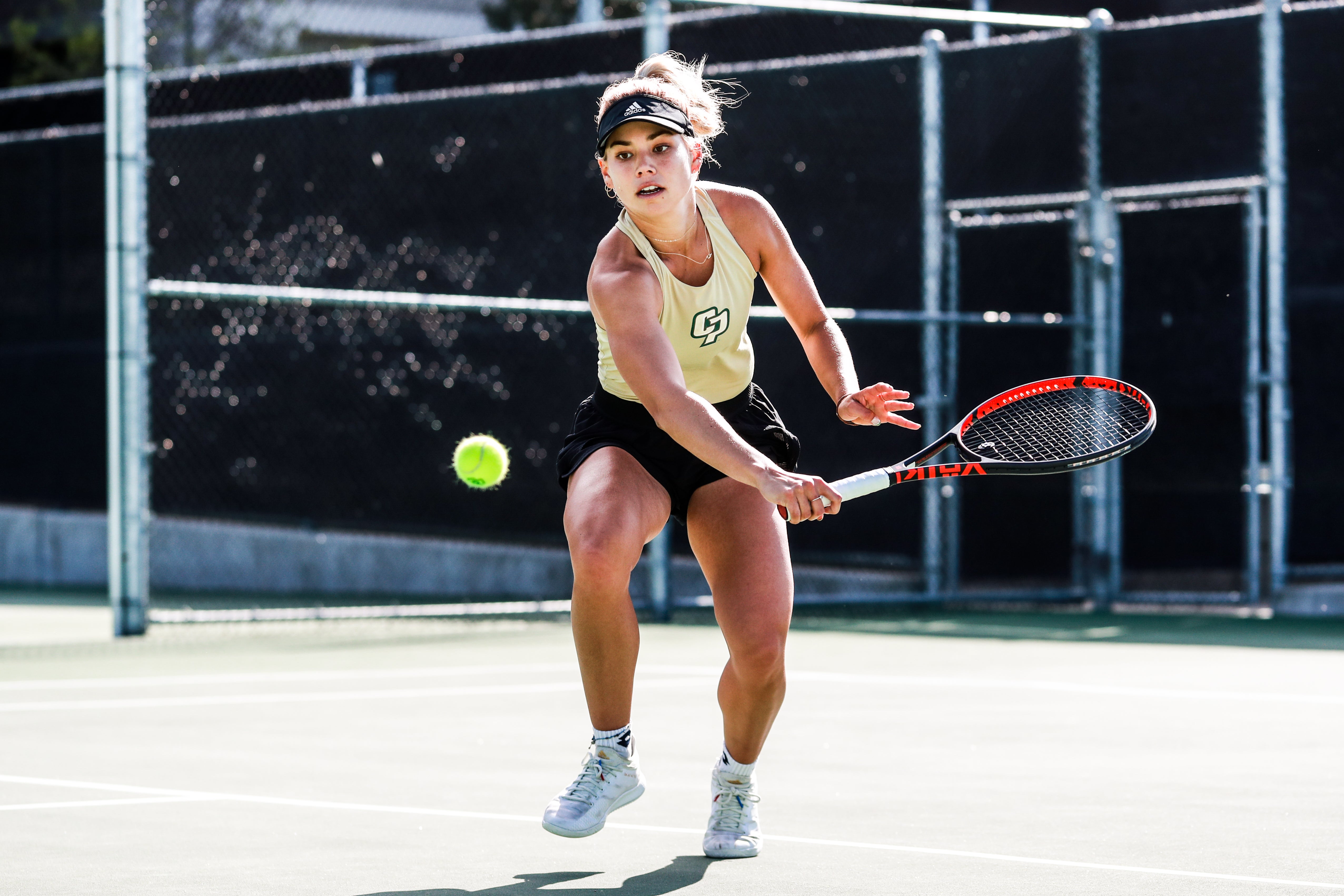 A woman wearing a Cal Poly tennis uniform hits a ball with her racket on the court.