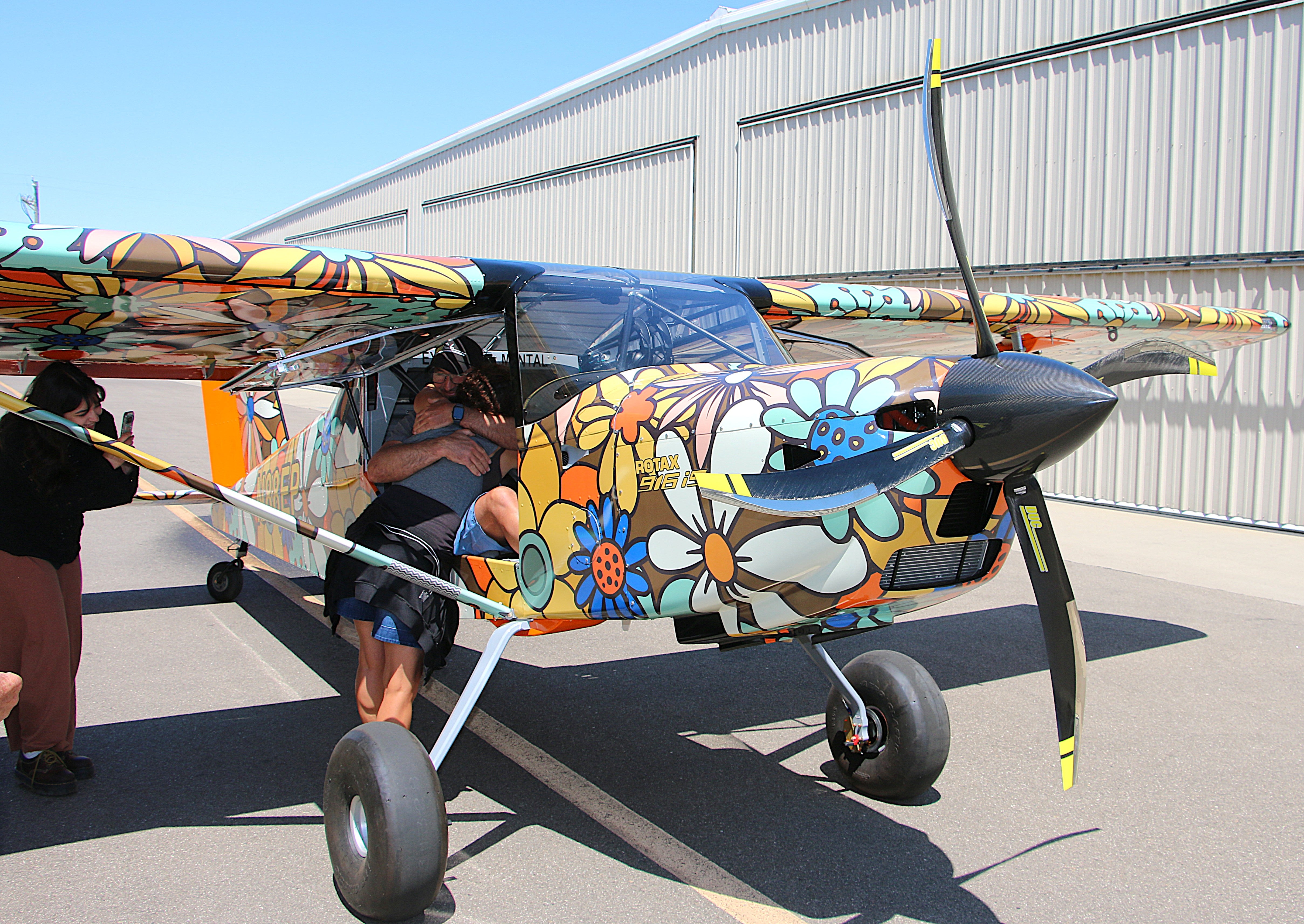 A woman hugs a man inside a floral patterned aircraft outside of an airplane hangar.