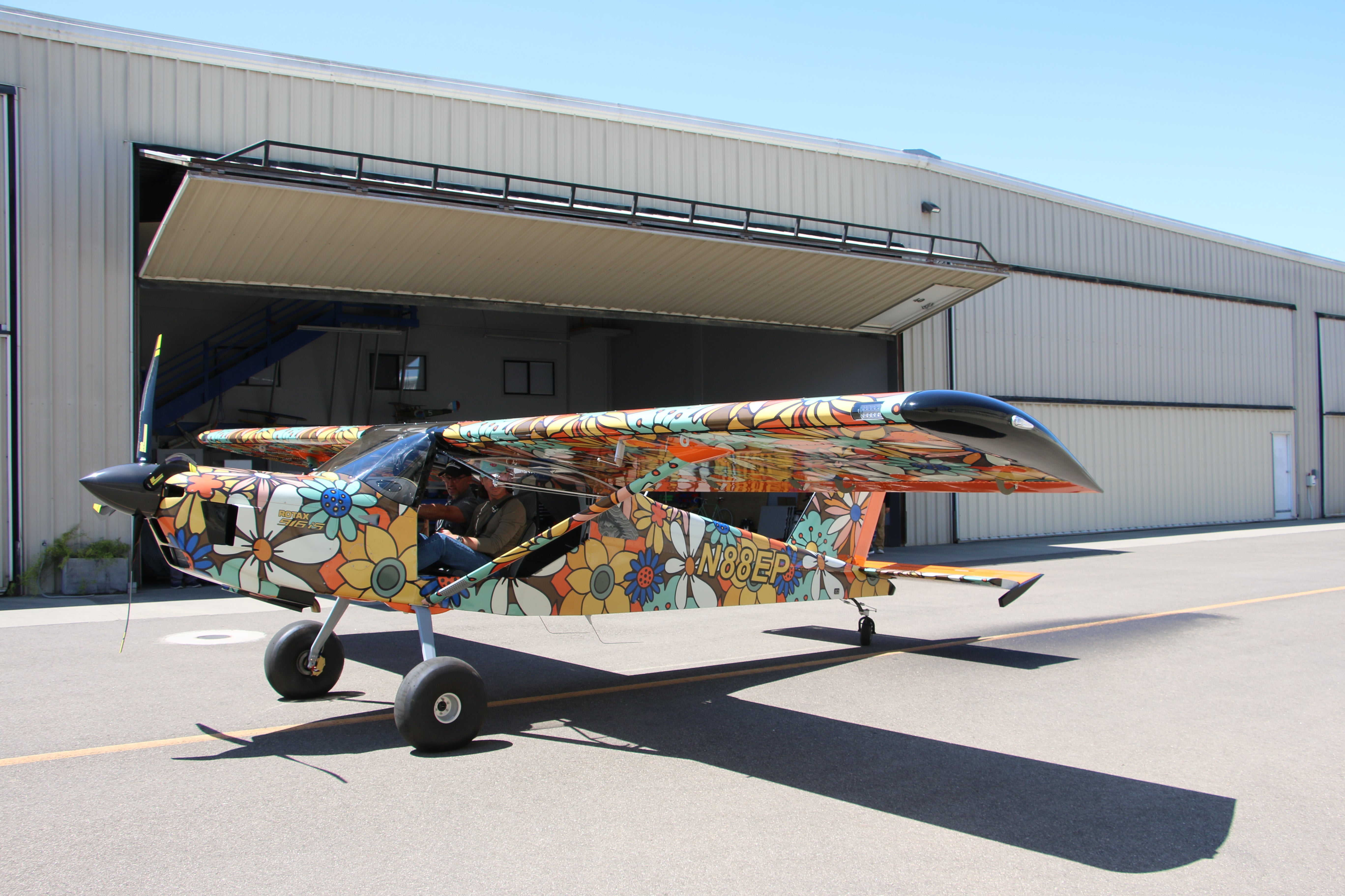 Two men sit in a floral-patterned airplane, taxiing out of a hangar at the airport.