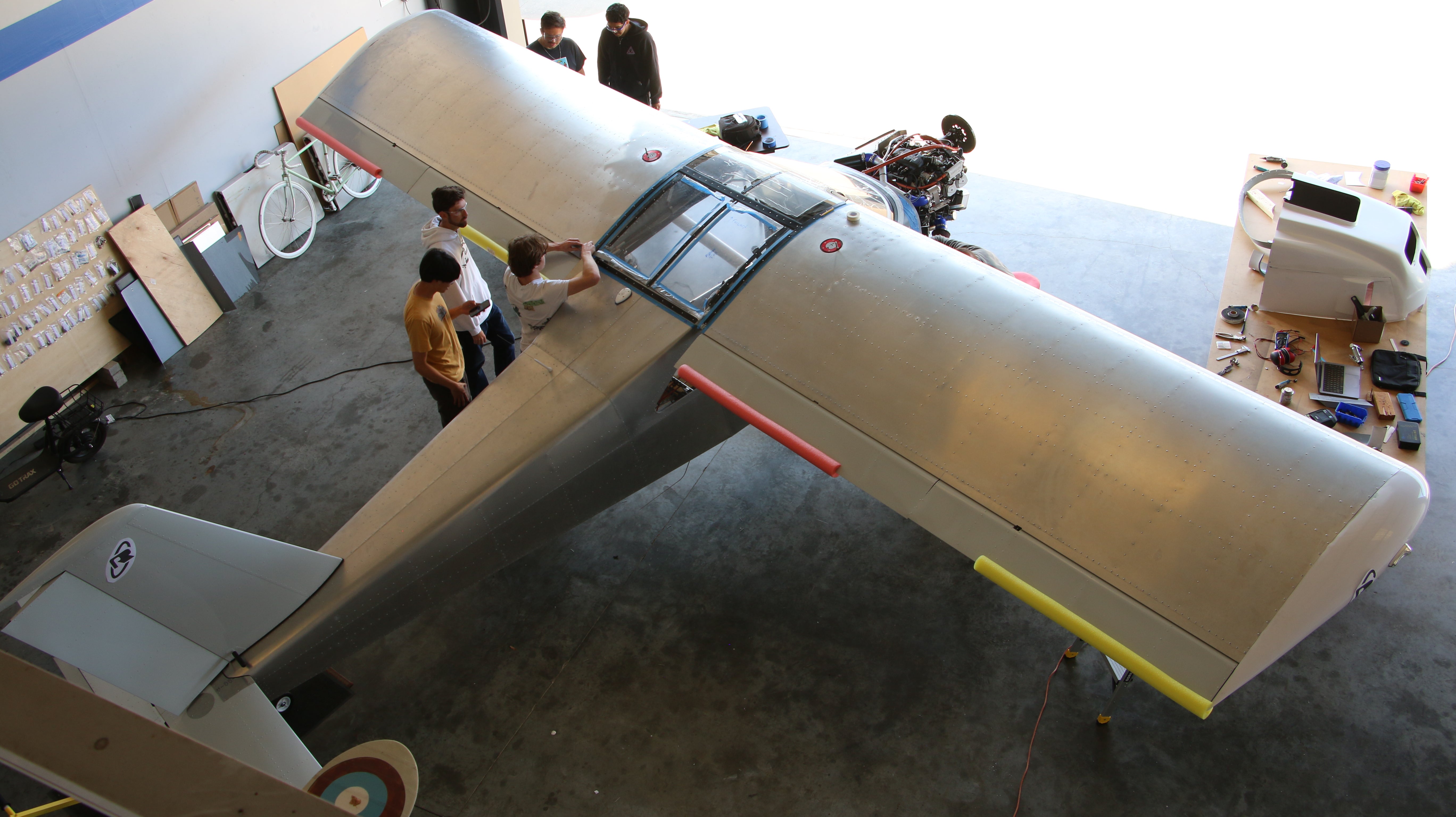 Students gather around a metallic aircraft in an airport hangar.