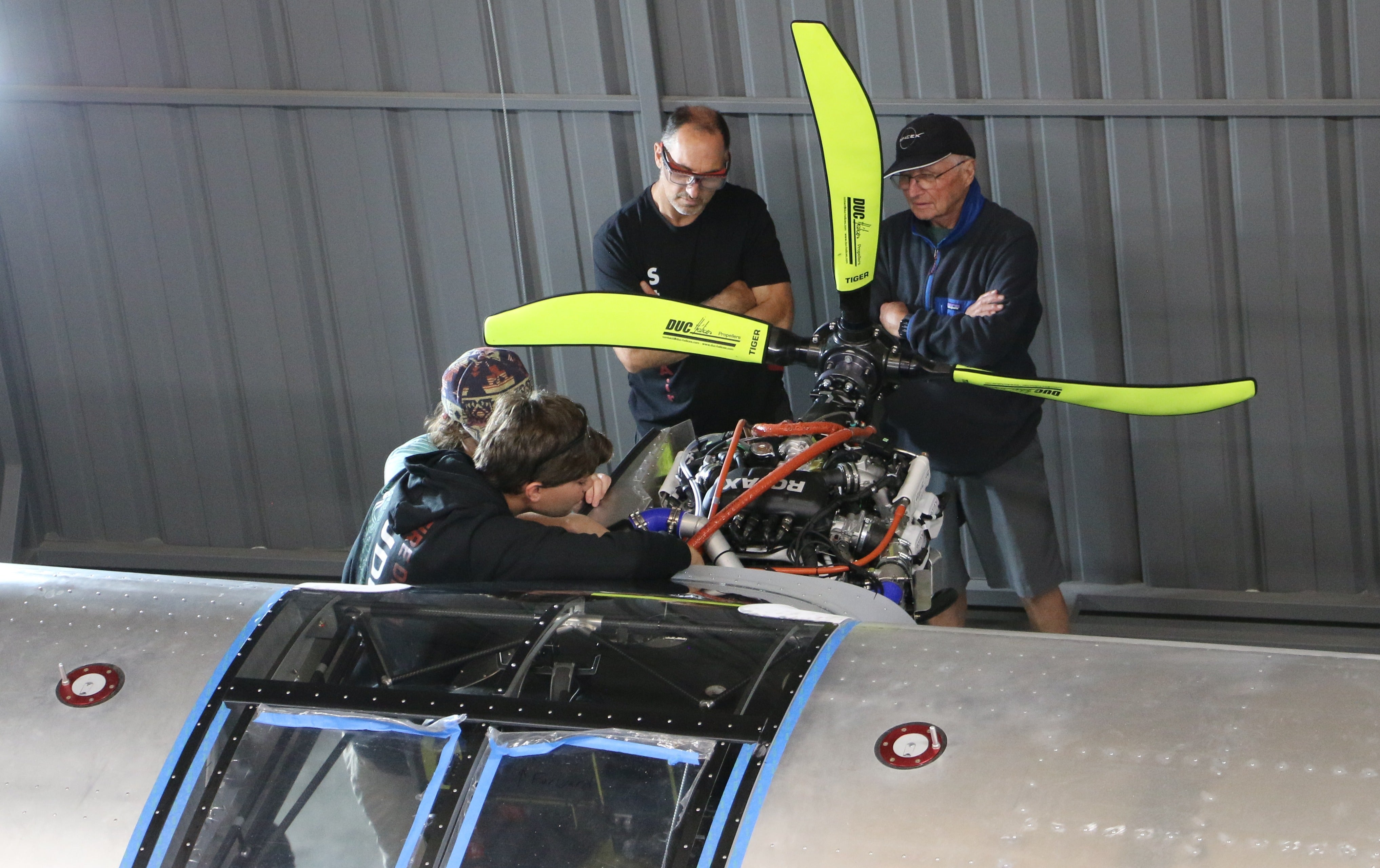 Two students attach the plane's propeller while the professor and his father look on.