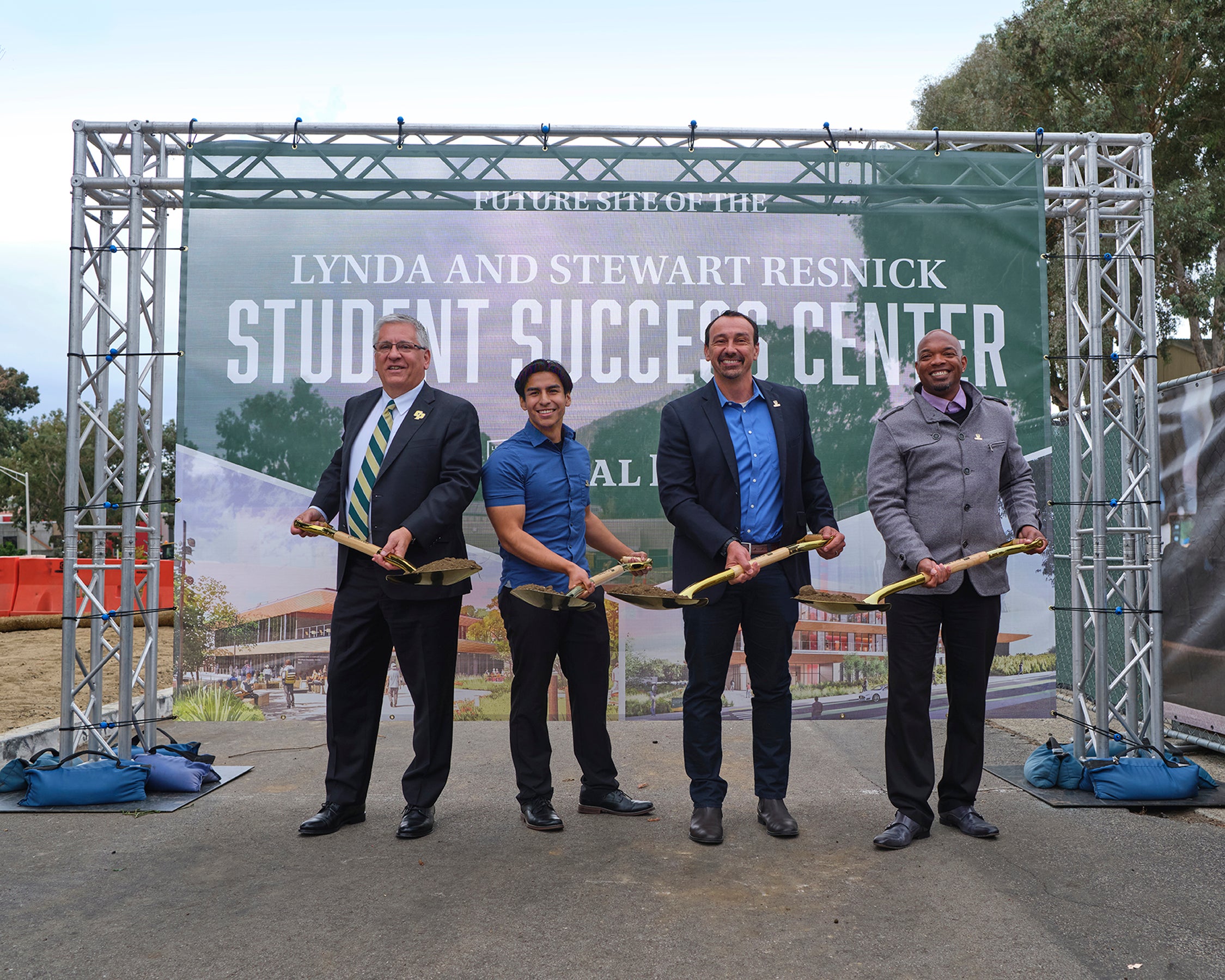 Four men, including Cal Poly President Jeffrey Armstrong, hold shovels and smile in front of a banner for the Lynda and Stewart Resnick Student Success Center.