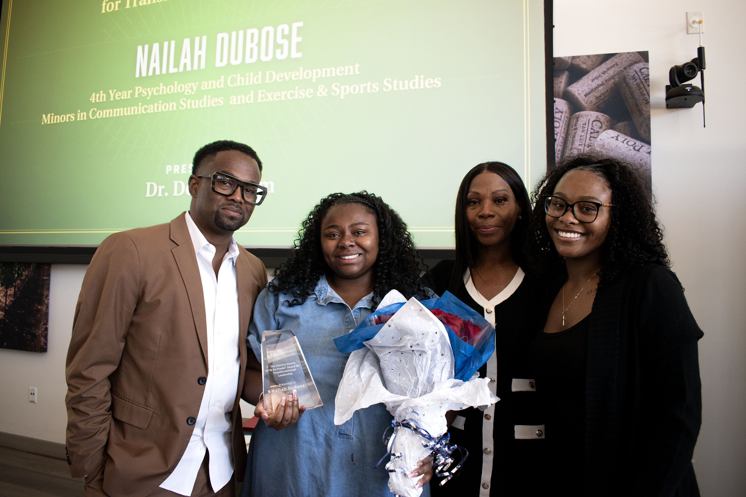 Nailah DuBose ('25) posing with her parents and sister during the President's Diversity Awards Ceremony 2025. The presentation screen behind them shows some of the words on the slide, "Nailah DuBose, 4th Year Psychology and Child Development, Minors in Communication Studies and Exercise and Sports Studies."