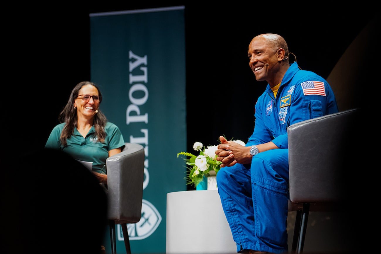 A woman in a green Cal Poly polo and a man in a blue jumpsuit sit in chairs onstage in front of a Cal Poly banner