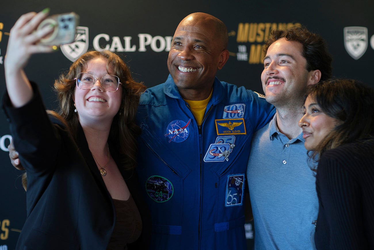 A man in a blue NASA jumpsuit smiles for a selfie with three college students