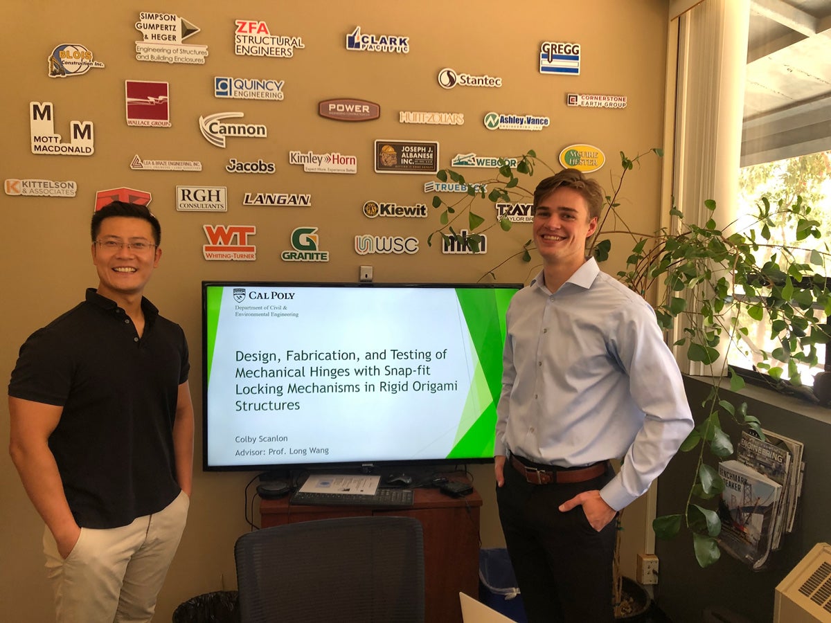 A professor and student stand next to a tv screen displaying a powerpoint presentation on their research into mechanical hinges.