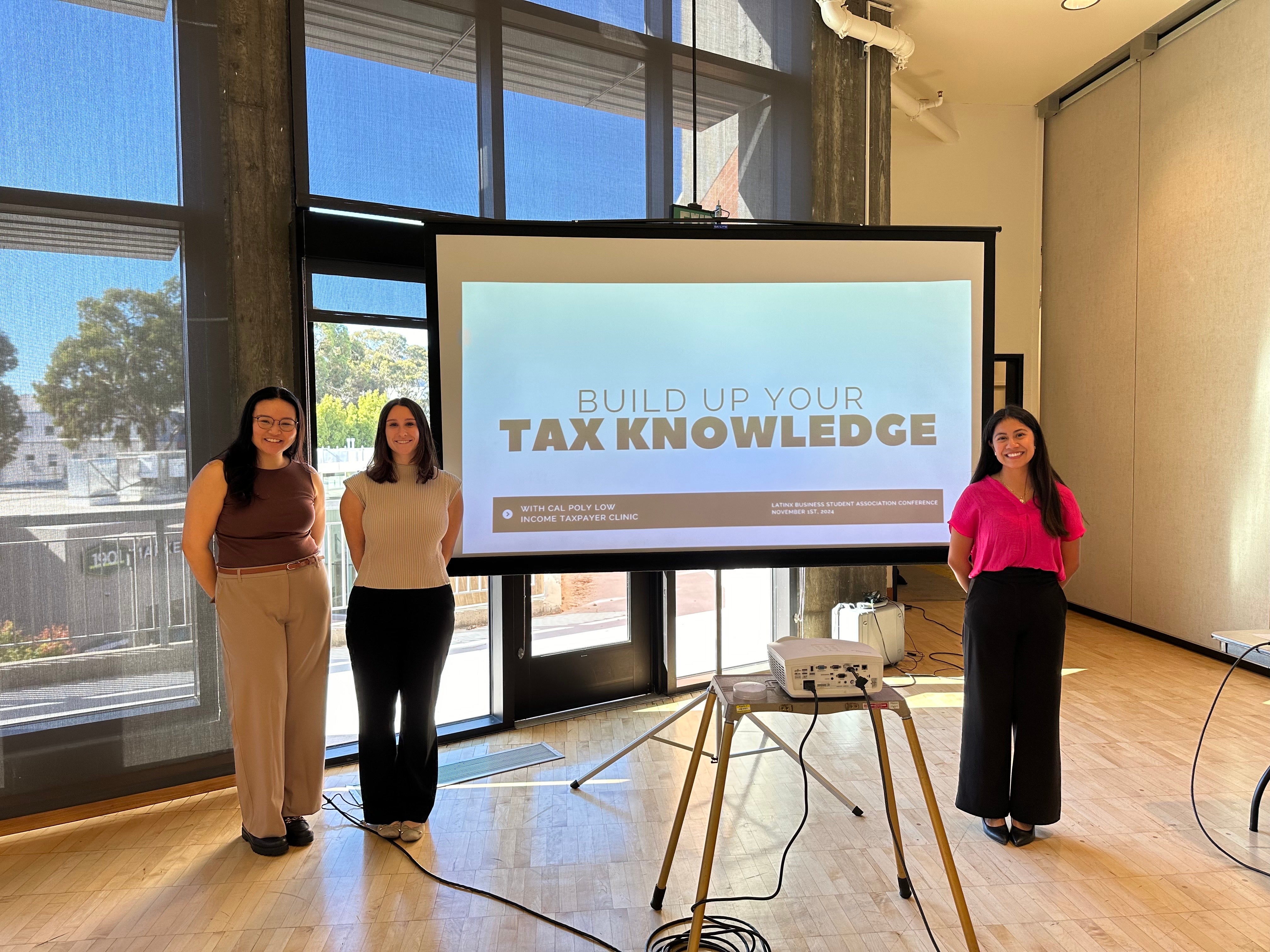 Three students stand next to a screen showing the start of a powerpoint that reads, Build up Your Tax Knowledge.