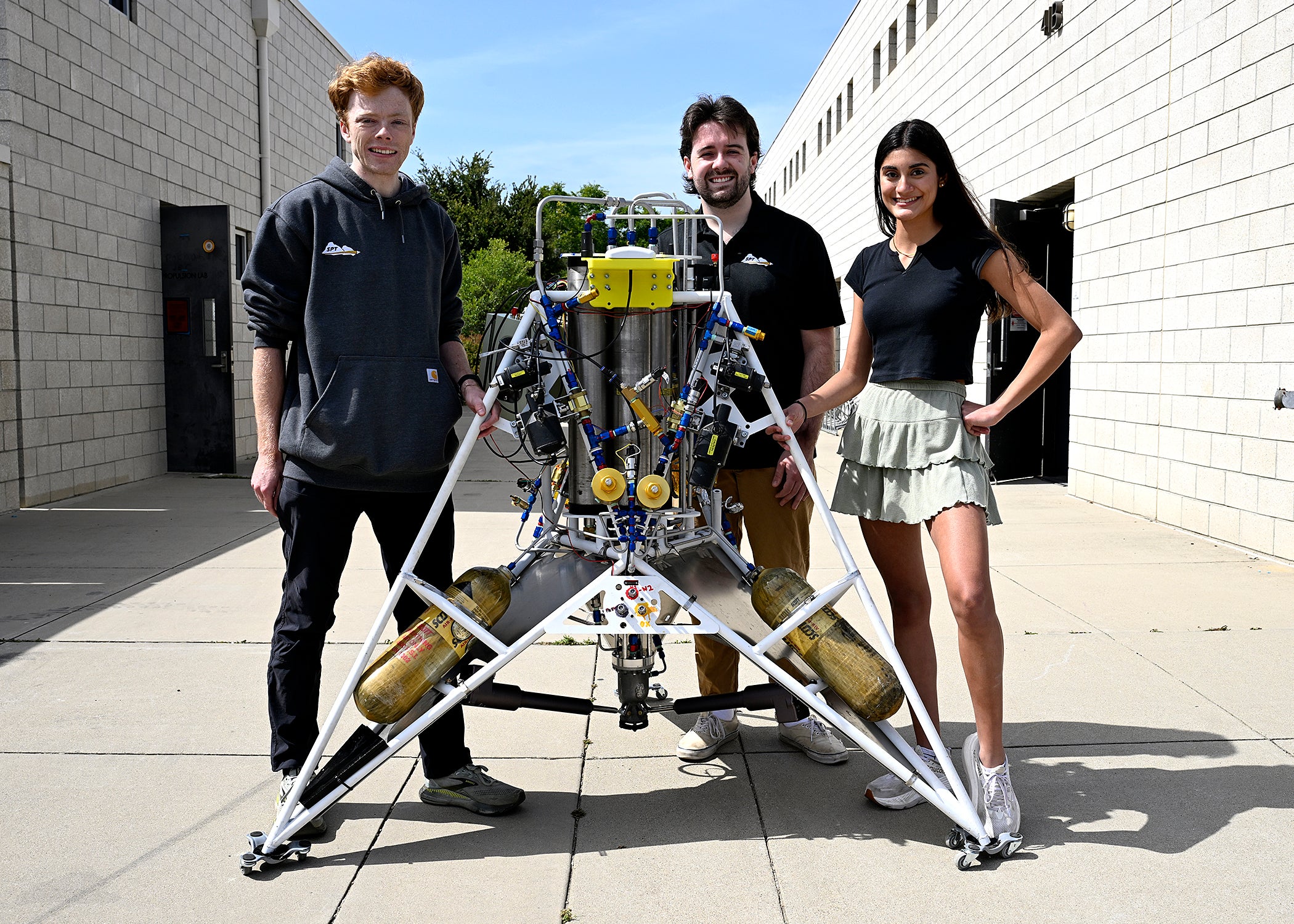 Three students pose outside the engineering building with a rocket machine.