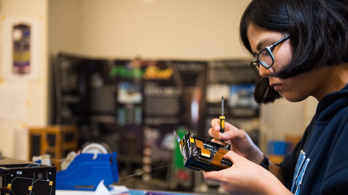 Student holds a screwdriver and works on a Cube Sat device.