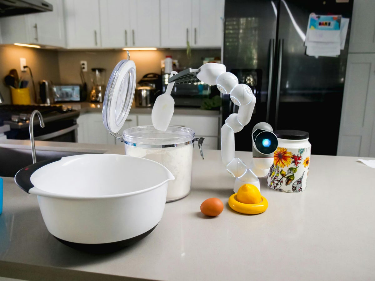 A robotic arm scoops flour from a canister into a mixing bowl in a kitchen.
