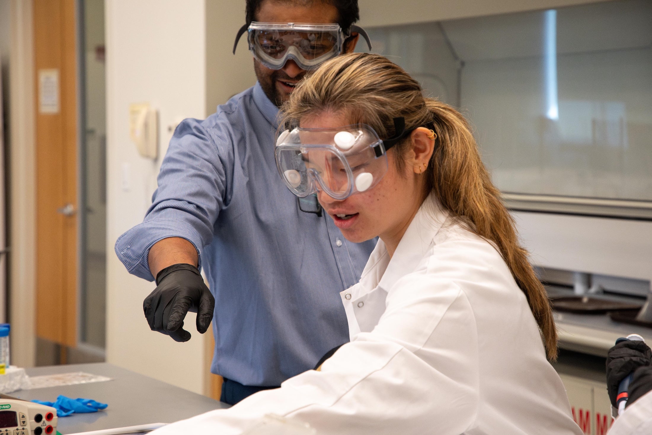 A professor and student wearing lab goggles conduct work.