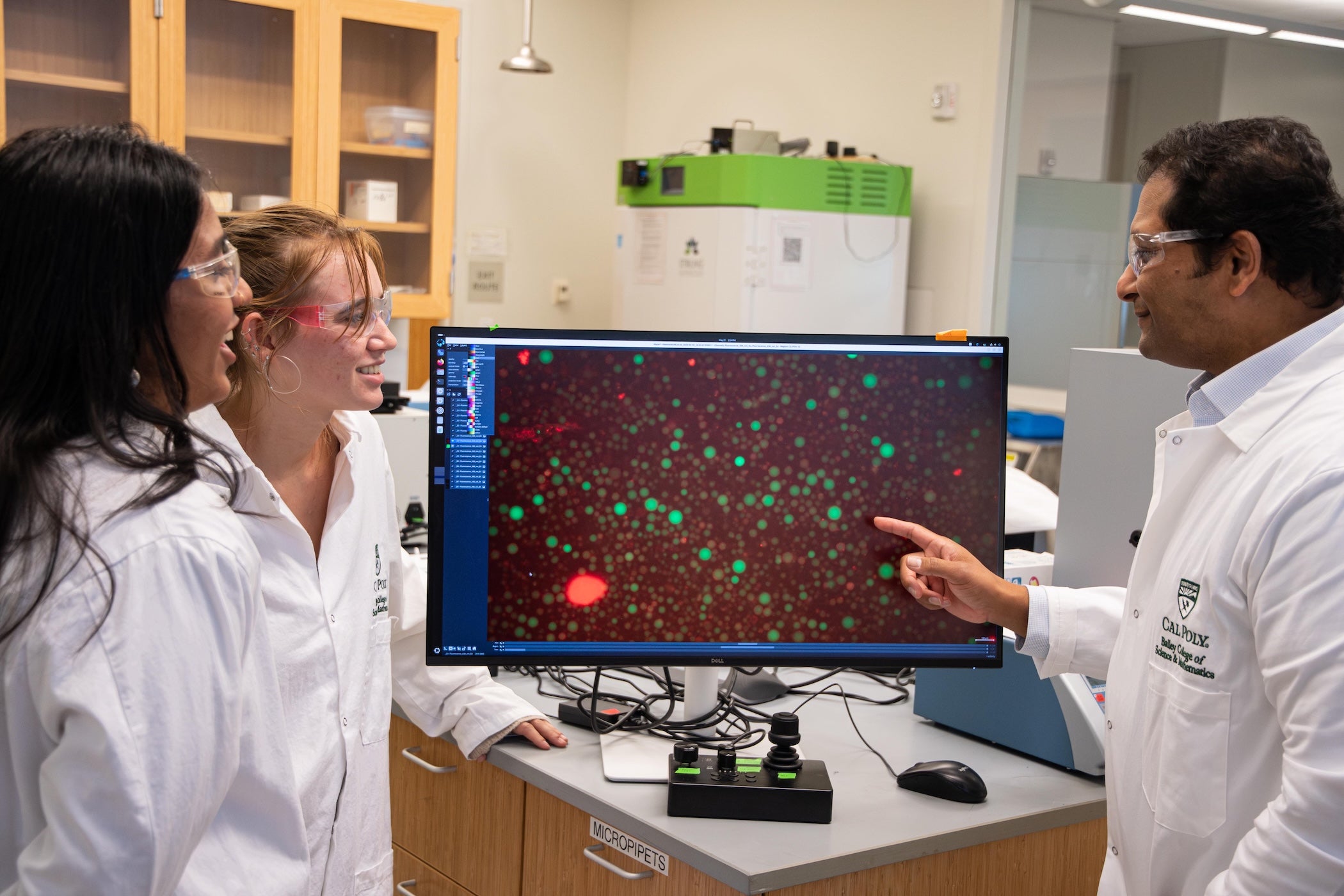 Two students and a professor in lab coats look at a model on a monitor.