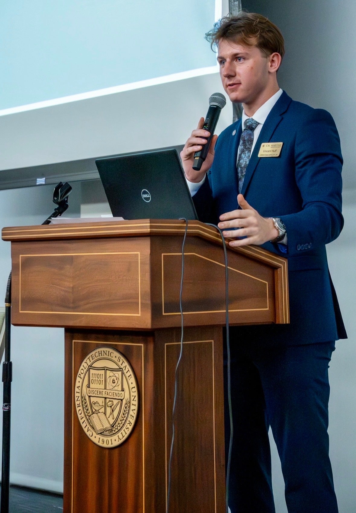 A student in a navy suit speaks at a wooden podium.