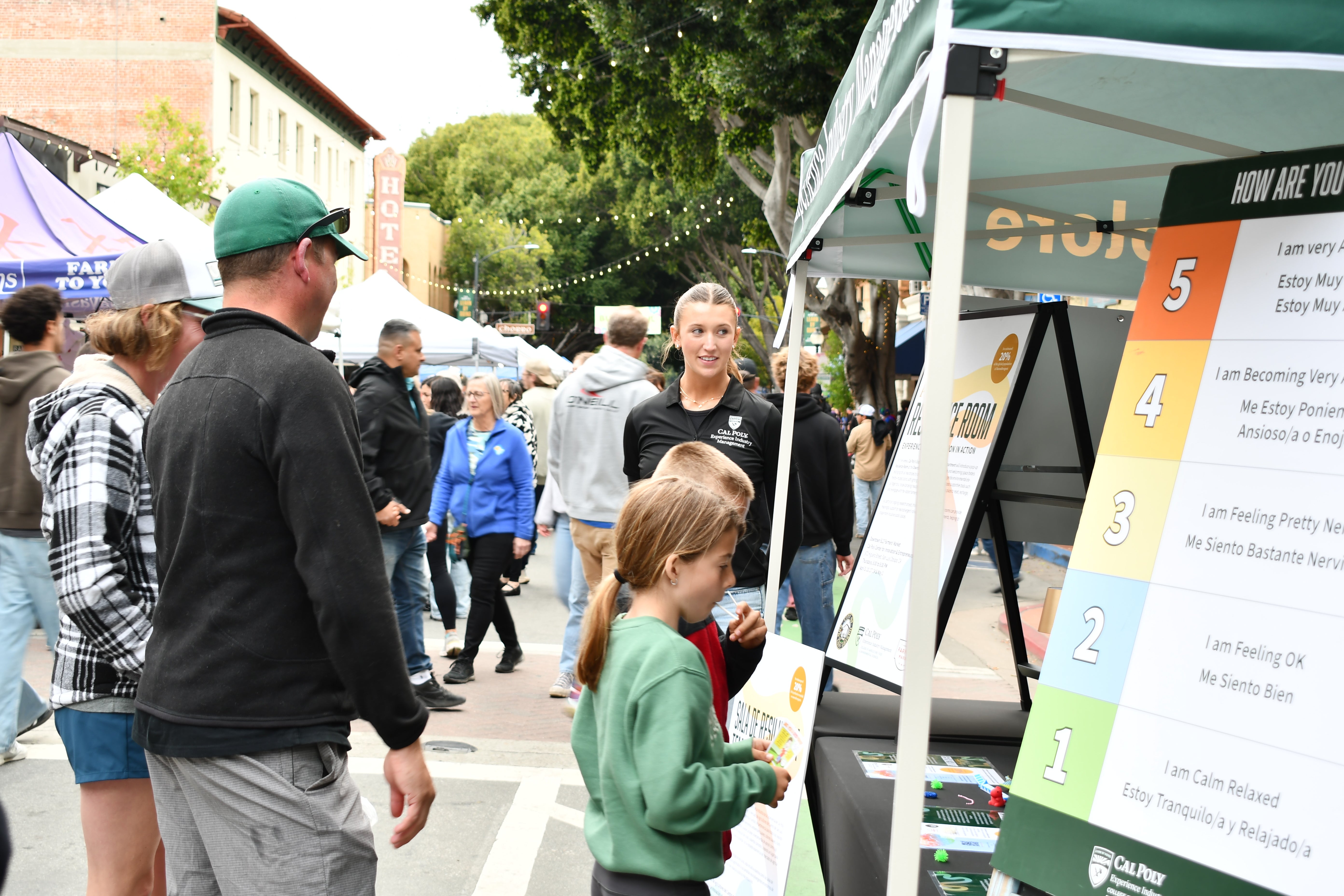A student at a farmers market booth speaks with adults and children passing by.