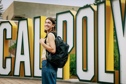 A student with a backpack standing in front of the Cal Poly letters sign.