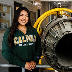 Denis Gonzalez-Reyes wearing a Cal Poly sweatshirt and standing next to a jet engine exhaust nozzle.