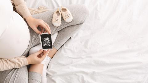 A pregnant person holding a sonogram with a pair of baby shoes on their leg.