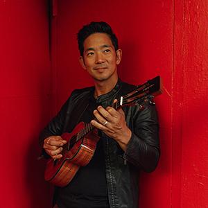 Jake Shimabukuro holding his ukulele and leaning on a red wall.