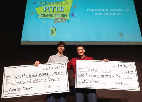 Two Cal Poly students standing on a stage holding large checks for $500 and $1,000 for winning the 2024 Elevator Pitch Competition.