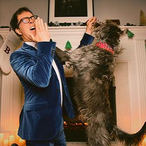 Ben Folds and a dog standing in front of a fireplace that’s been decorated for the holidays.