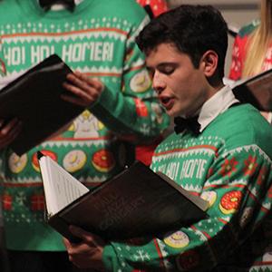 A closeup of a student wearing a holiday sweater and caroling with a choir in the background.