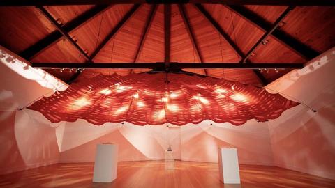 A wood-and-fabric structure hanging from the ceiling in an art gallery.