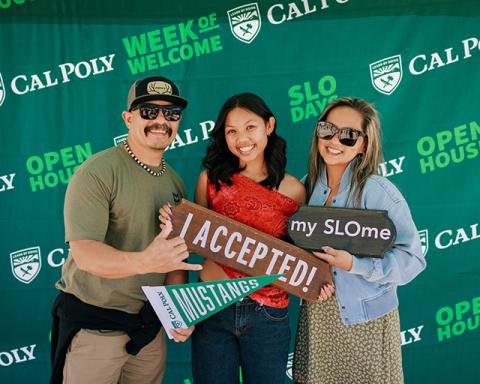 A student standing with her parents in front of a Cal Poly banner holding a pennant and signs that say, “I accepted!” and “my SLOme.”