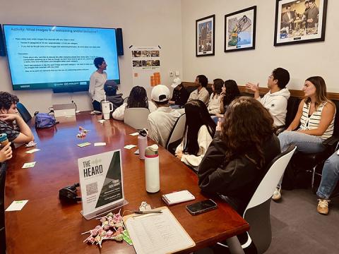 Students at a conference table provide feedback to a speaker in front of a Powerpoint presentation.