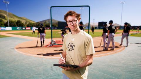 Gabe Riedel holding a clipboard and standing on the field of Baggett Stadium with the baseball team in the background.