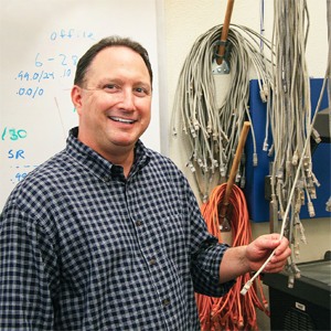 Hugh Smith standing in a room on campus and holding an ethernet cable with a whiteboard in the background.