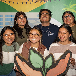Members of a cultural club standing in front of an ASI backdrop, holding a sign in the shape of hands holding a plant.