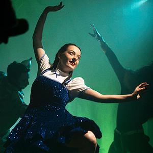 A ballerina dressed up as Dorothy dancing on stage with the Tin Man and the Cowardly Lion dancing in the background.
