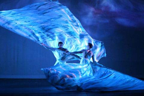 Dancers performing on stage with blue lighting and sheets of fabric dangling around them.