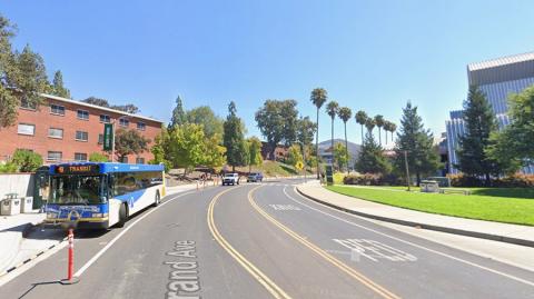 A bus parked along Grand Avenue with the red brick dorms and Performing Arts Center in the background.