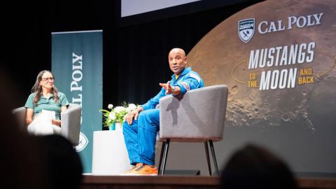 Victor Glover talking about the Artemis II mission on stage with a banner in the background showing the moon and the Cal Poly logo.